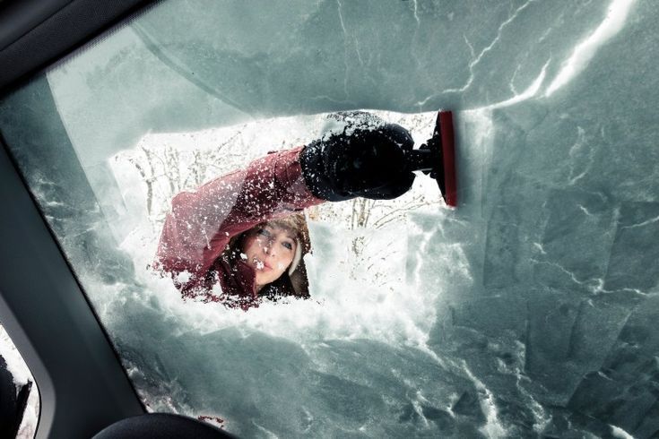A woman scraping ice off her car windshield on a winter morning, dressed in a red jacket, with snow 