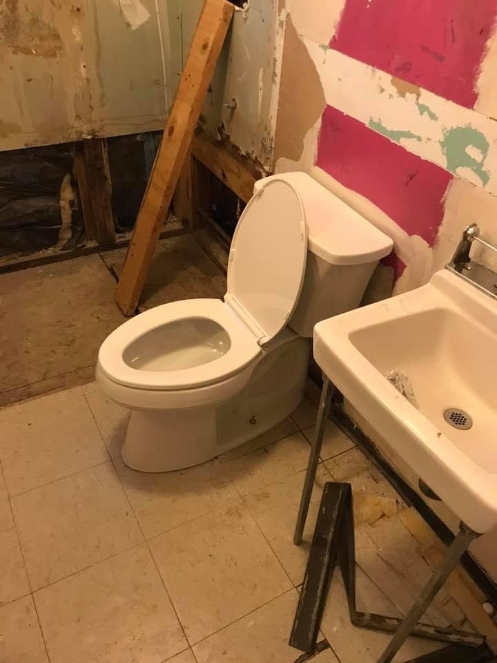 Small bathroom with an open toilet, sink, scattered tools, and peeling painted walls.