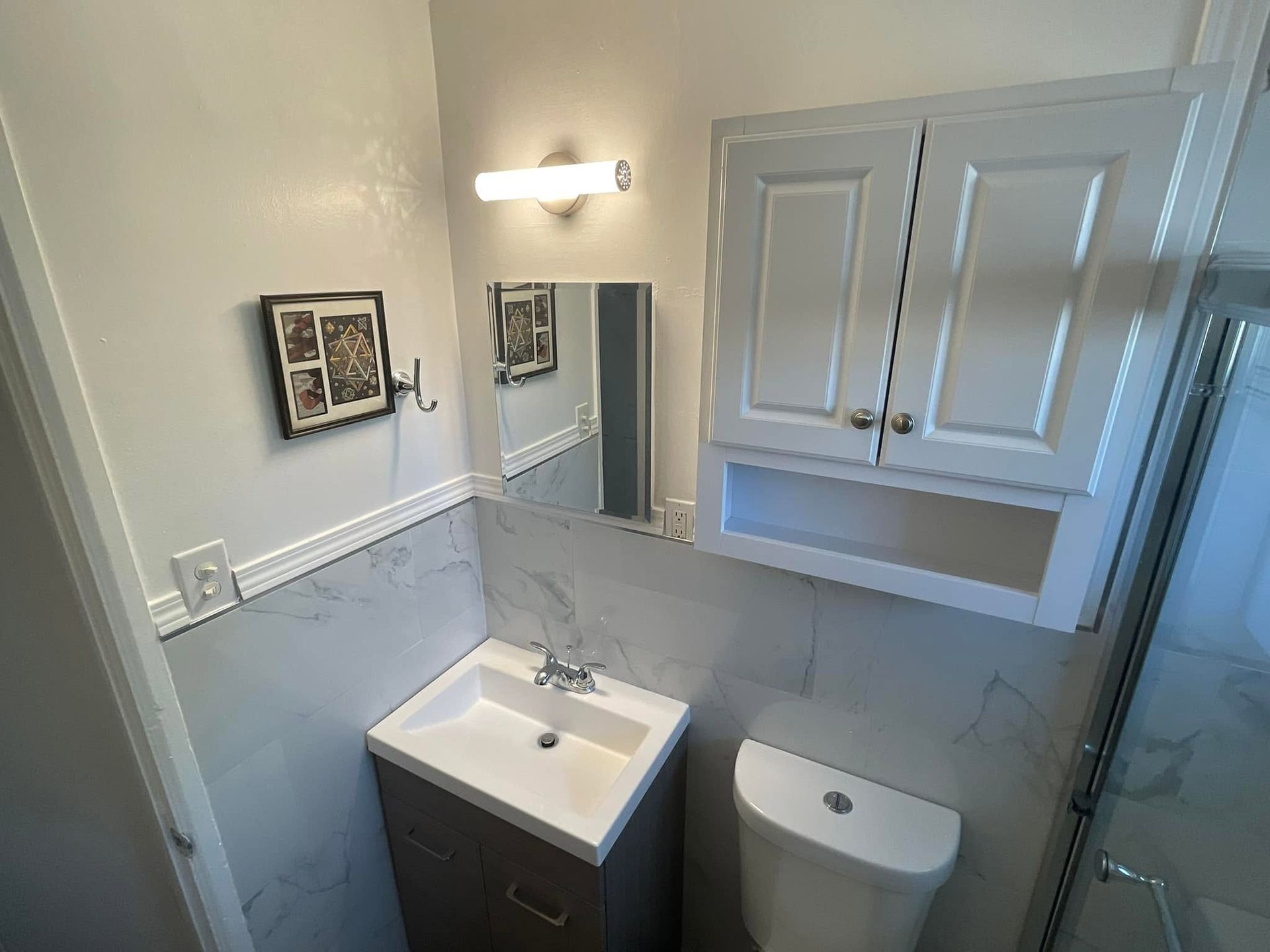 Small bathroom with white cabinets, pedestal sink, toilet, mirror, and glass shower door.