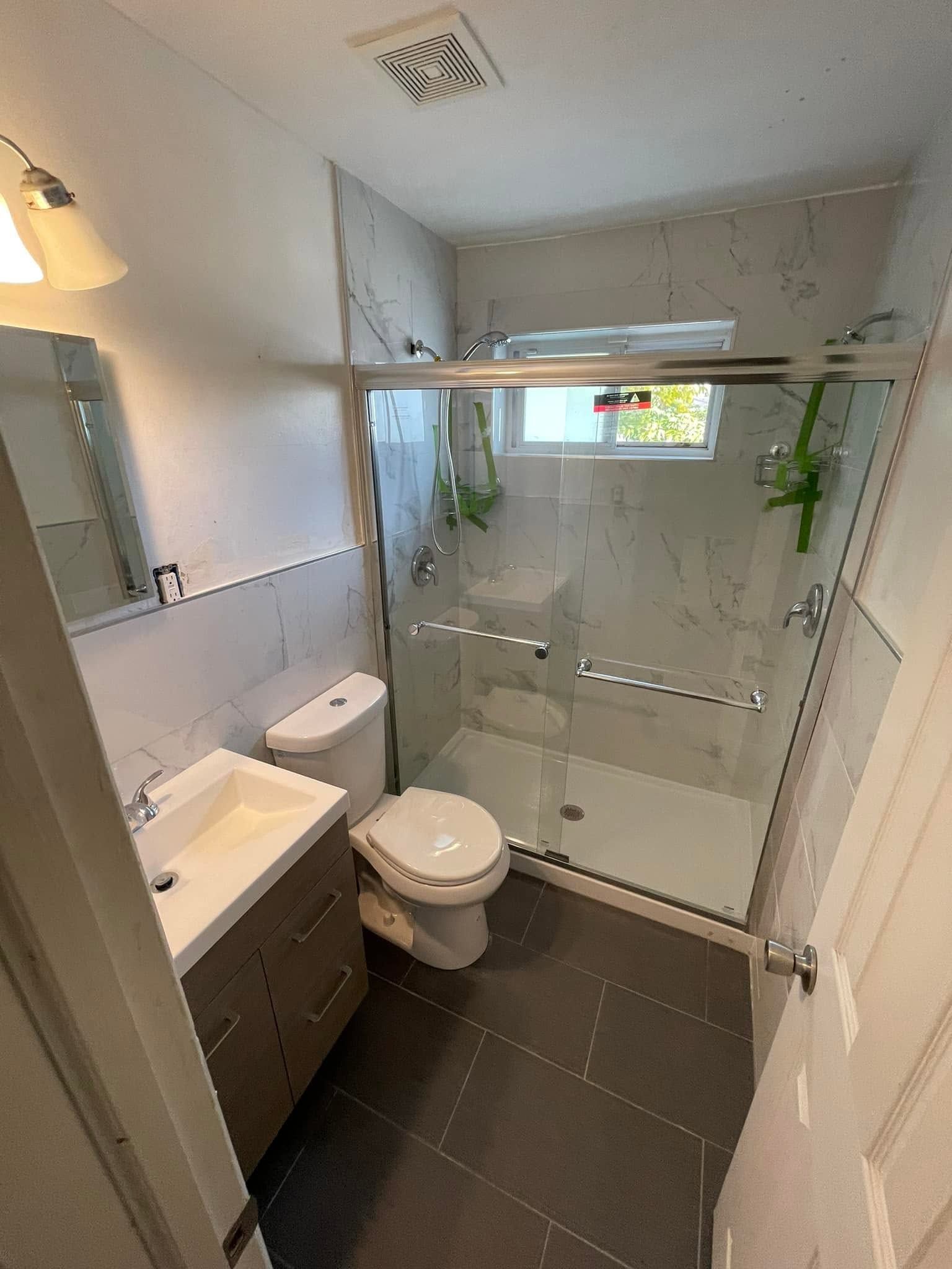 Small modern bathroom with shower, toilet, sink, and gray tile floor, viewed from the doorway