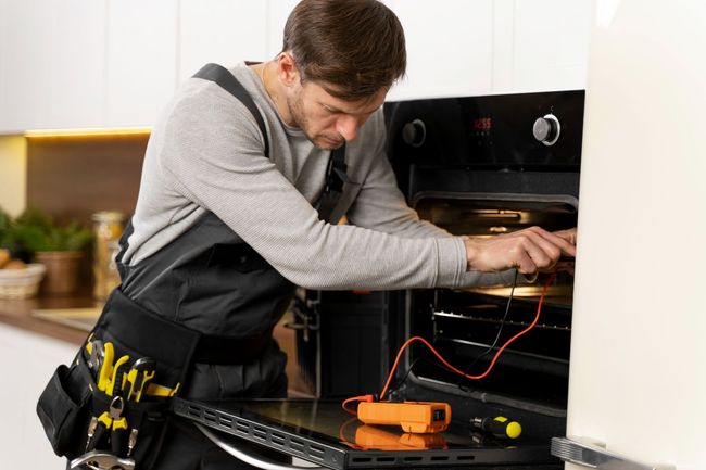 Electrician repairing an oven with a multimeter in a kitchen setting.