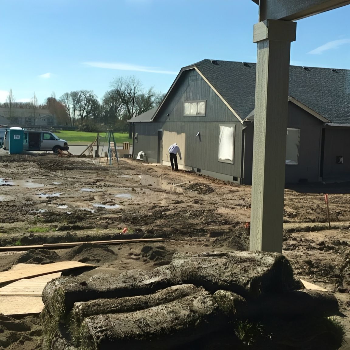 New home under construction with muddy yard, gray siding, and a worker near the house