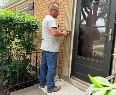 Man standing at a glass patio door beside a brick house, holding the handle and looking inside