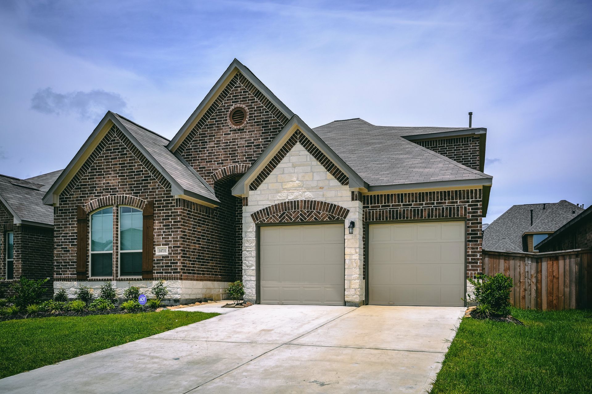 Suburban brick house with two white garage doors, front lawn, and concrete driveway under a cloudy sky