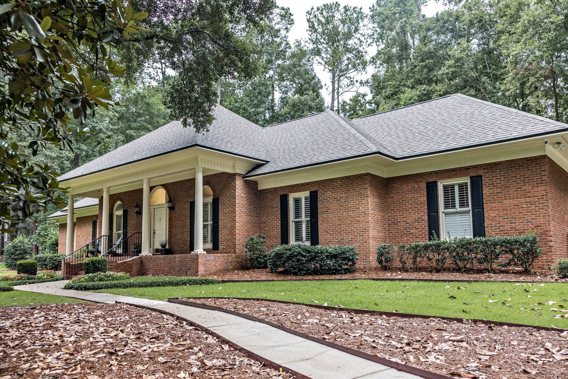 Brick suburban house with gray roof, front porch, and green lawn surrounded by trees