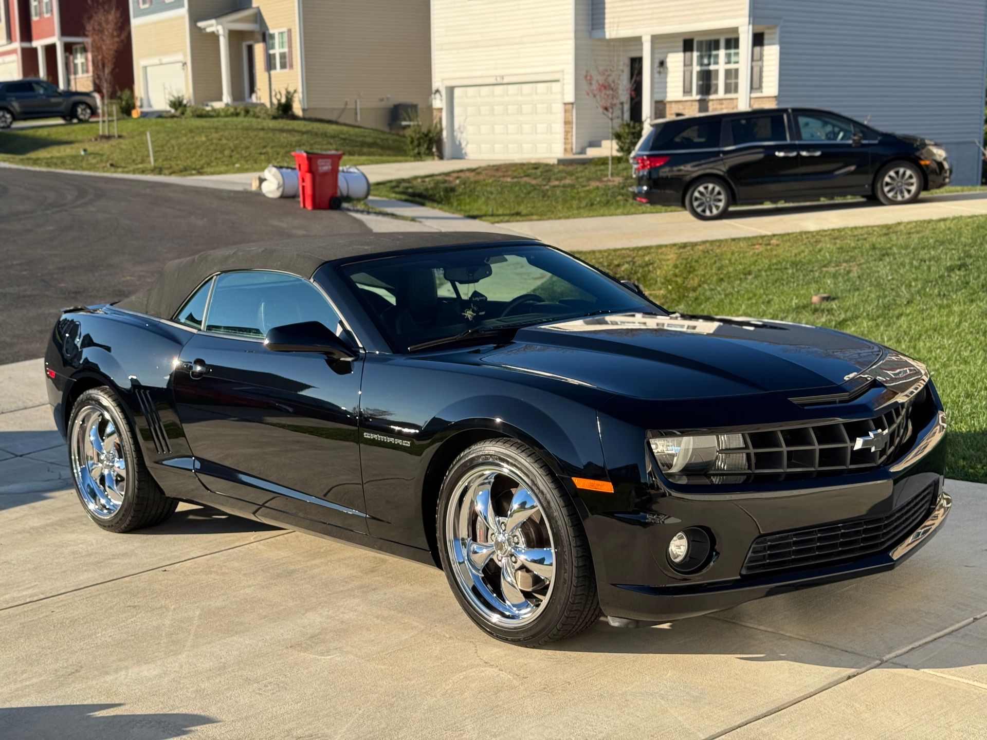 Black convertible Chevrolet Camaro parked on a driveway in front of houses; shiny chrome wheels.
