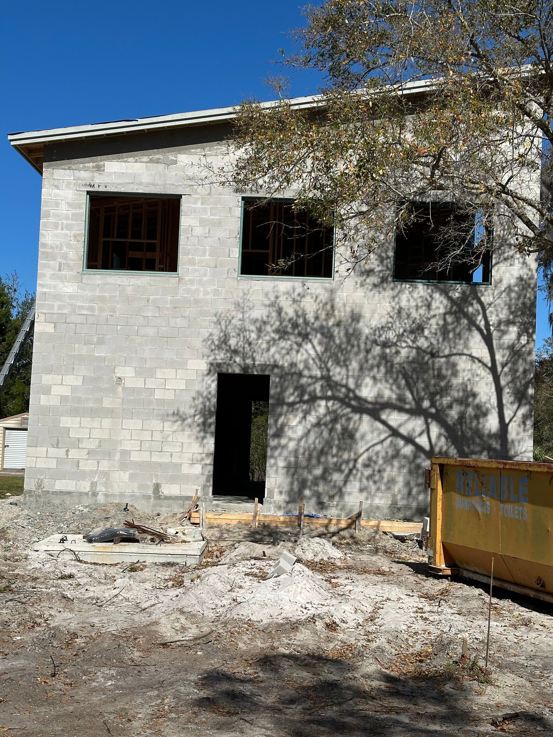 Two-story white cinderblock house under construction with open window and door, tree shadows, and a yellow dumpster