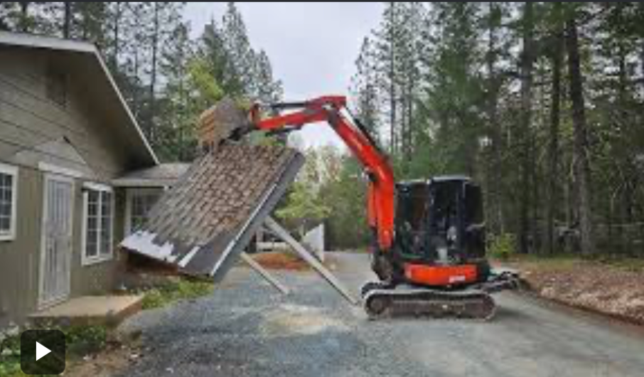 Orange excavator lifting a roof section beside a house in a wooded driveway