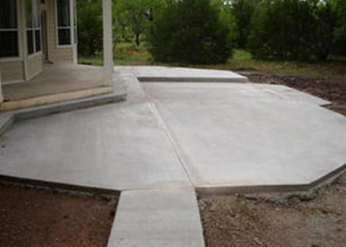 Concrete patio and walkway beside a house, with a raised step and surrounding dirt.