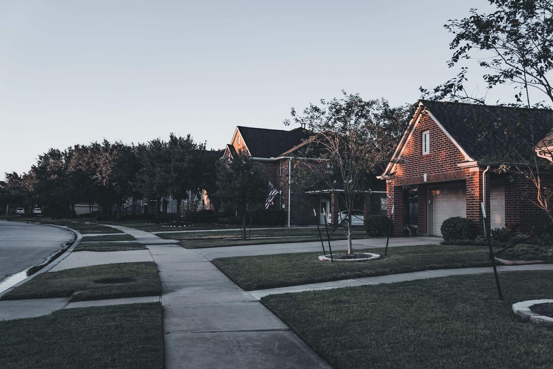 Suburban street at dusk with sidewalks, lawns, and houses lit by warm porch lights