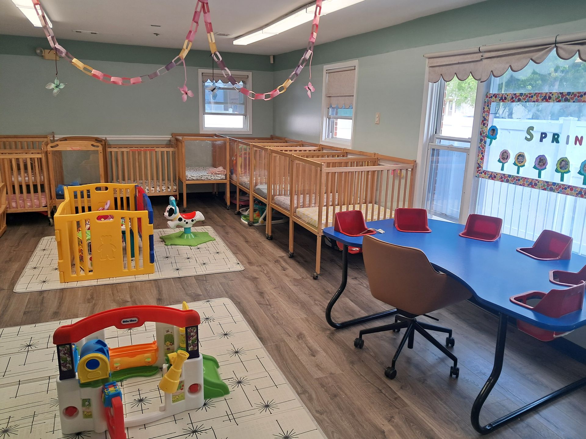 Daycare playroom with cribs, a blue table and chairs, toys, and spring decorations