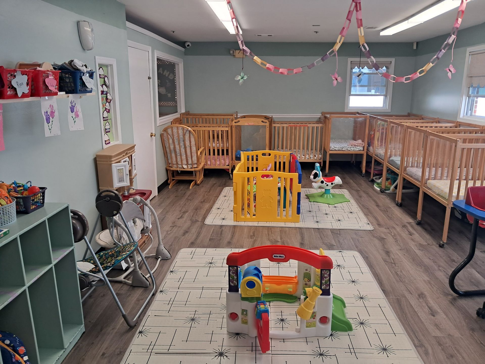 Childcare playroom with wooden cribs, colorful toys, and foam play mats on a wood floor