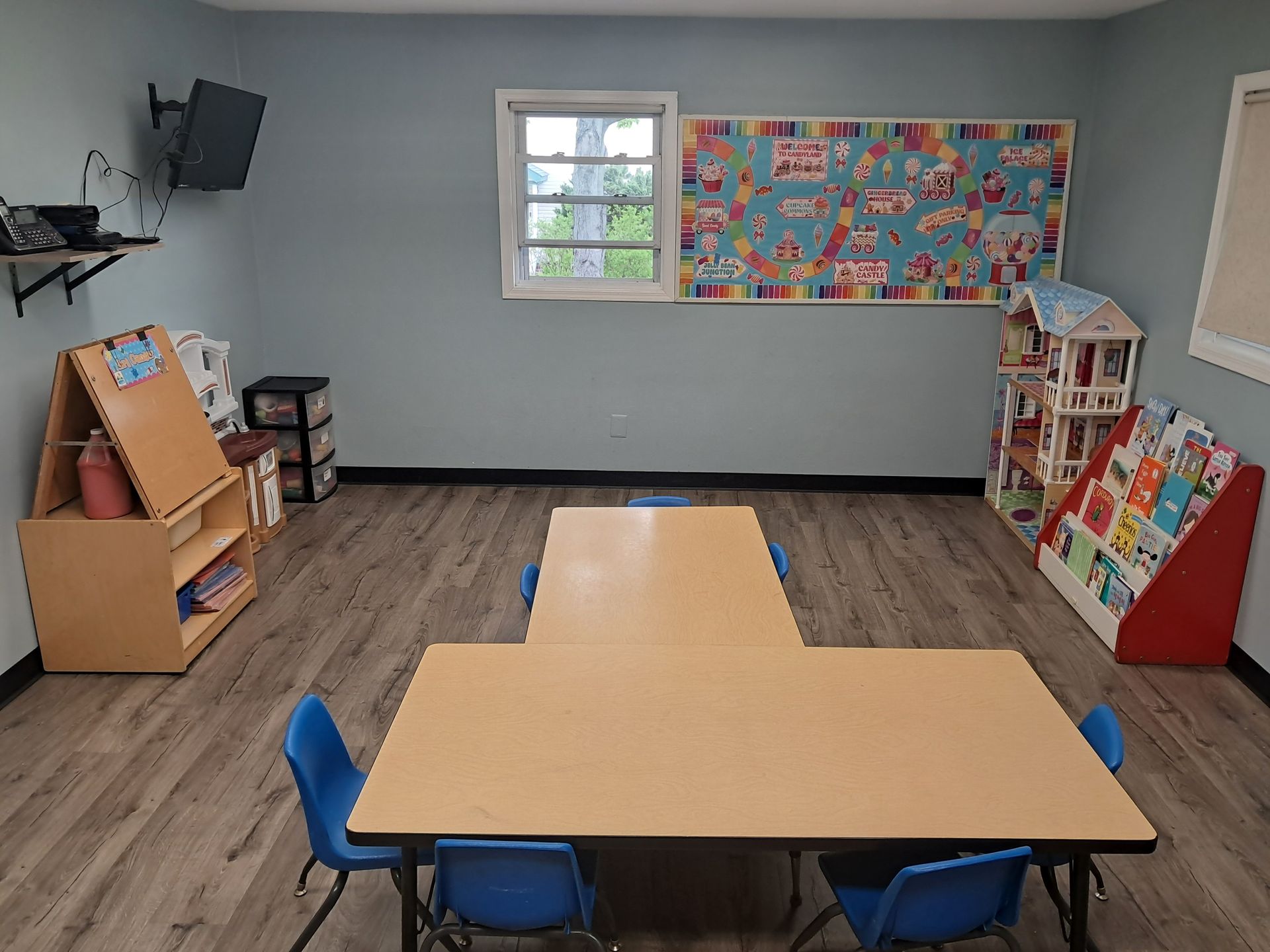 Empty classroom with wooden tables, blue chairs, bookshelves, and colorful bulletin boards.