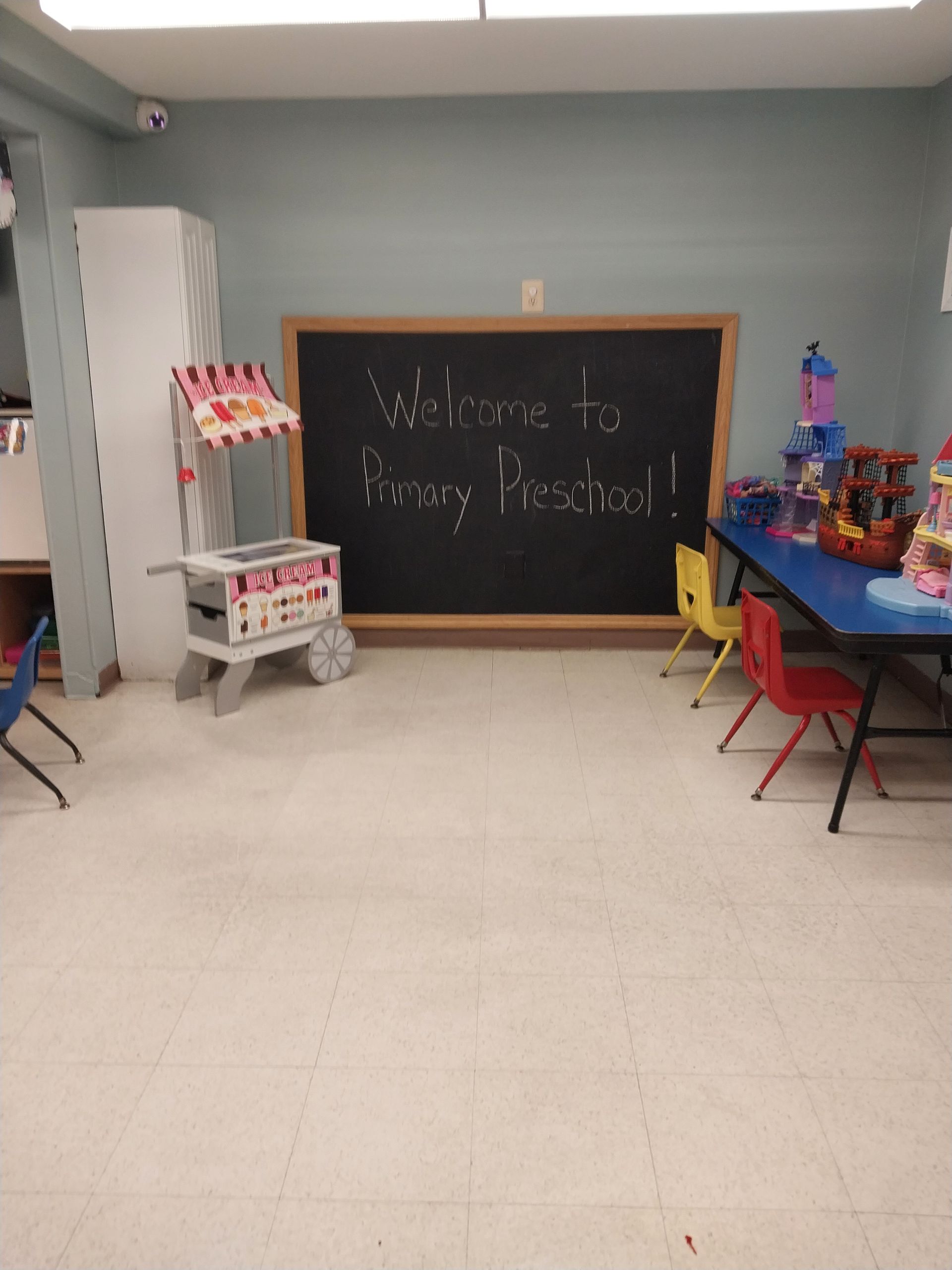 Classroom play area with a chalkboard reading “Welcome to my classroom,” tables, chairs, and toys