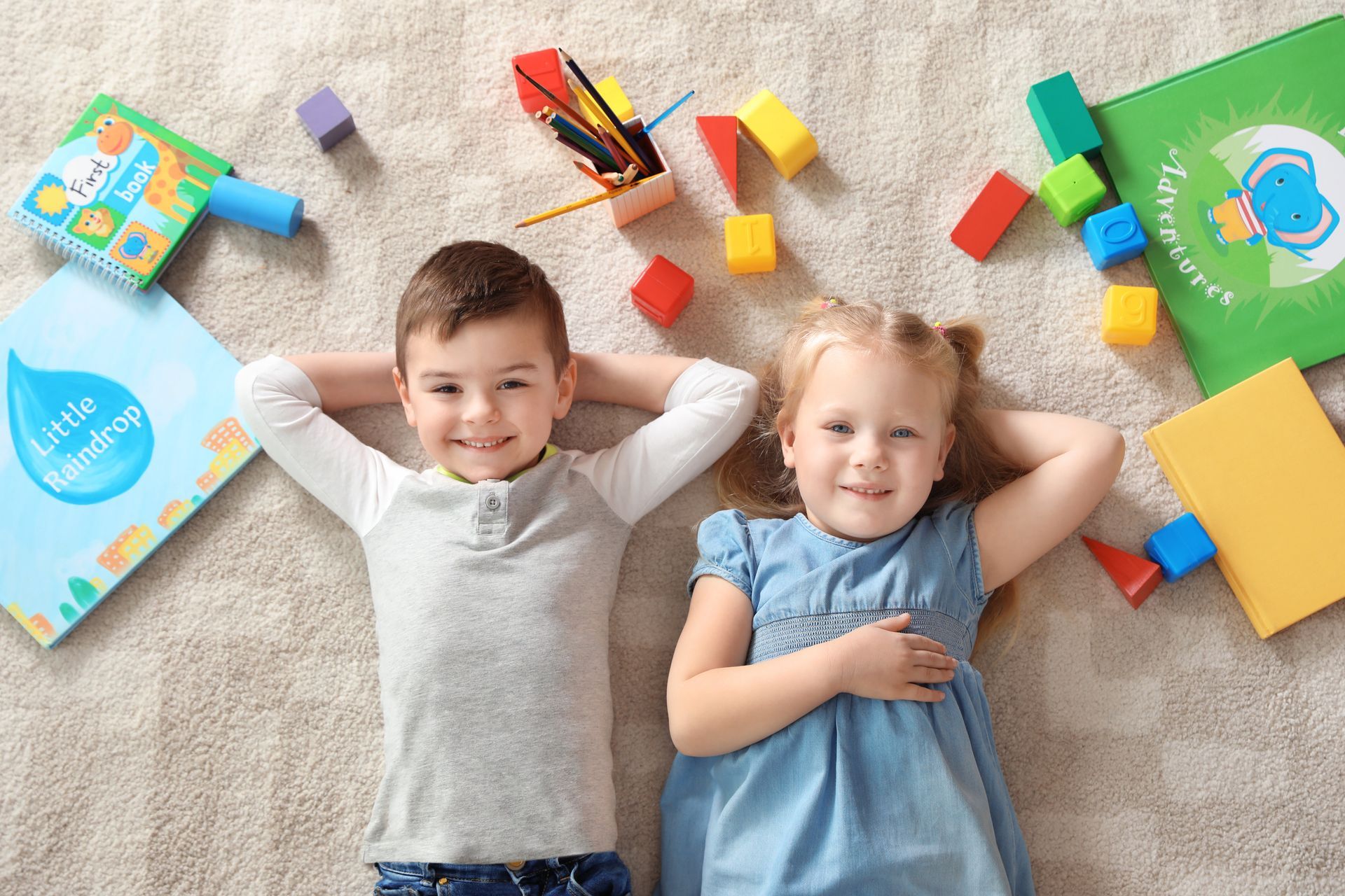 Two children lying on carpet, smiling among colorful building blocks and toys