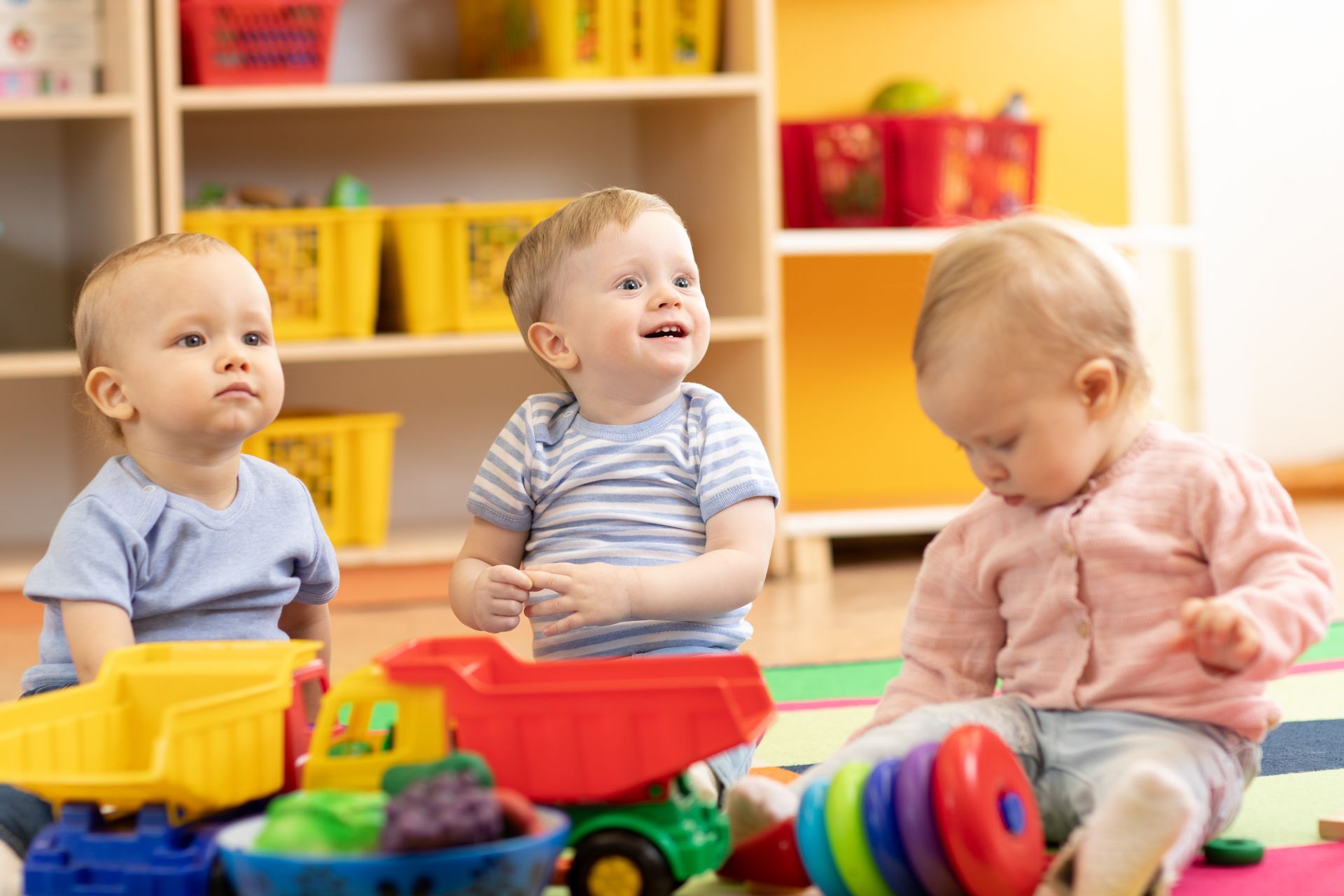 Three toddlers playing with colorful toys on the floor in a bright classroom
