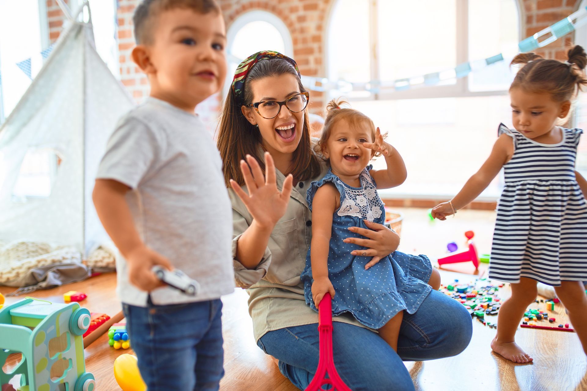 Adult and three children playing together in a bright indoor playroom, smiling and waving