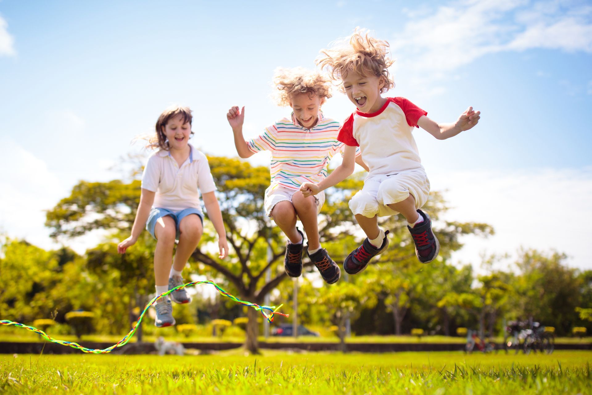 Three children jumping on a trampoline in a sunny park with green grass and trees