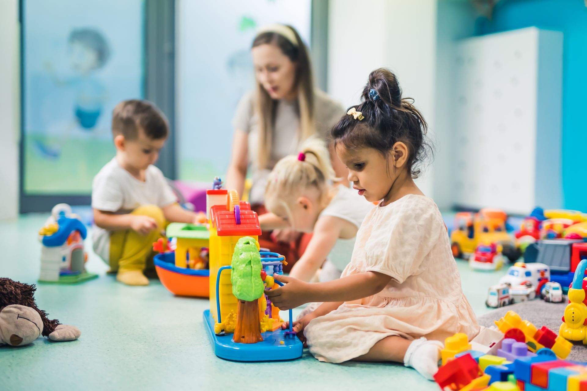 Children playing with colorful toys in a classroom while a teacher supervises.