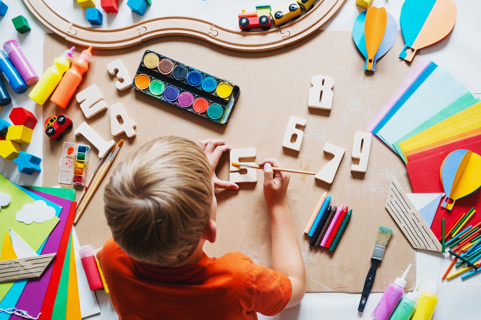 Child drawing with colorful art supplies on a table, with crayons, paper, and paint arranged around them