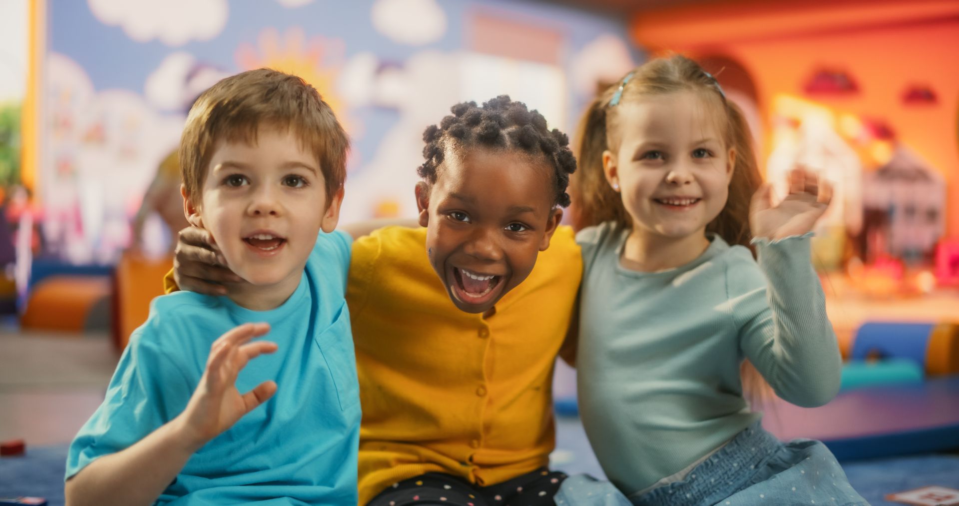 Three smiling children sit together on a couch in a colorful playroom.
