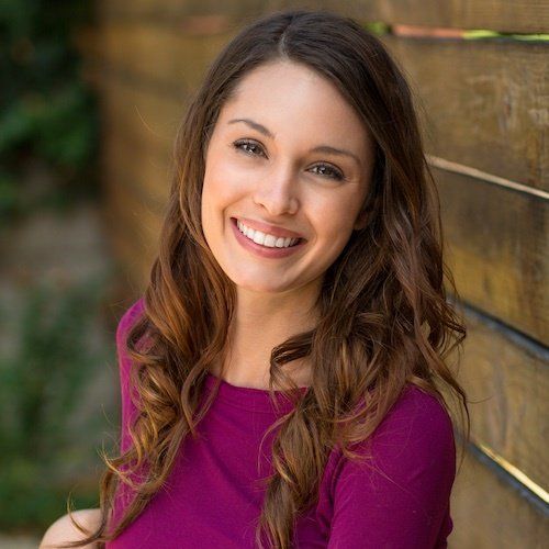 A woman in a purple shirt is smiling in front of a wooden fence.