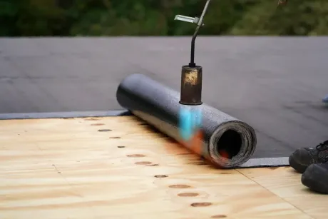 A worker using a torch to adhere roofing material to a wooden surface.