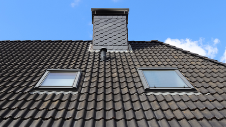 A roof with dark tiles, a chimney, and two skylights against a blue sky.