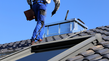 Roofer in blue suit on a tiled roof, working near a metal structure, bright blue sky.