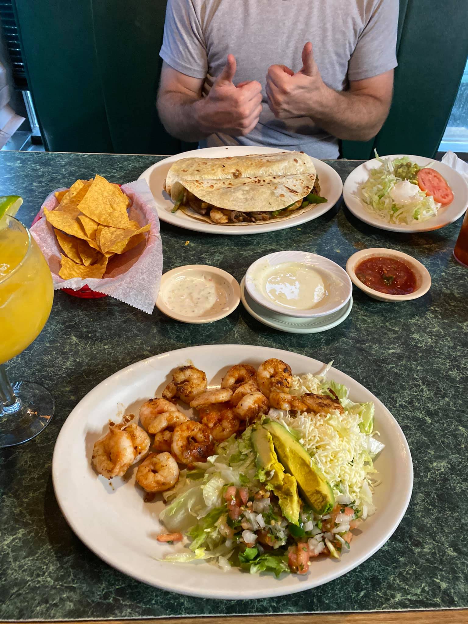 Table with Mexican food: shrimp, quesadilla, chips, salsa, and guacamole. Person gives thumbs up.