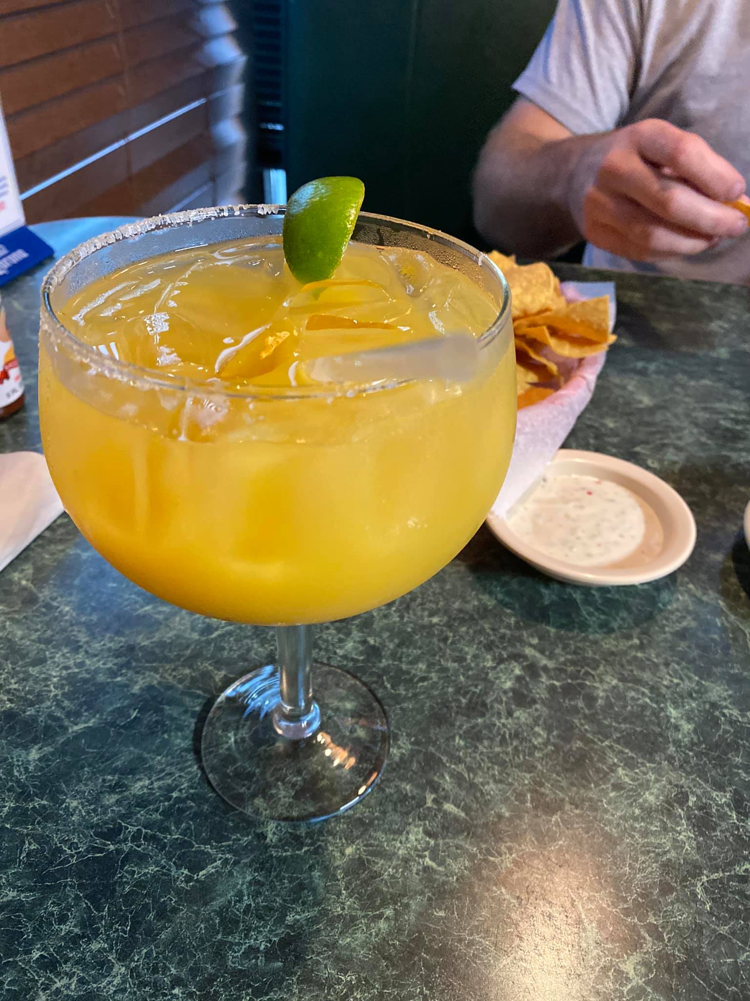 Mango margarita in a salt-rimmed glass with a lime wedge, next to chips, dip, and person.