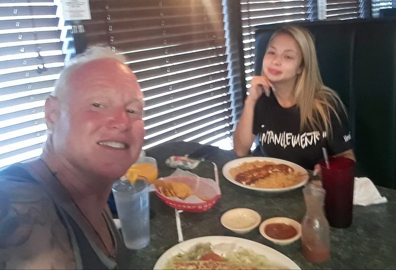 Two people at a restaurant table. Man smiling, taking selfie. Woman smiling, eating a meal. Food and drinks visible.