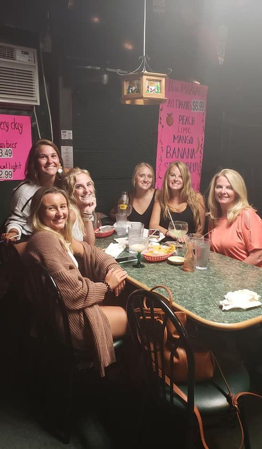 Six people sit at a table in a dimly lit bar, smiling. There are snacks and drinks.
