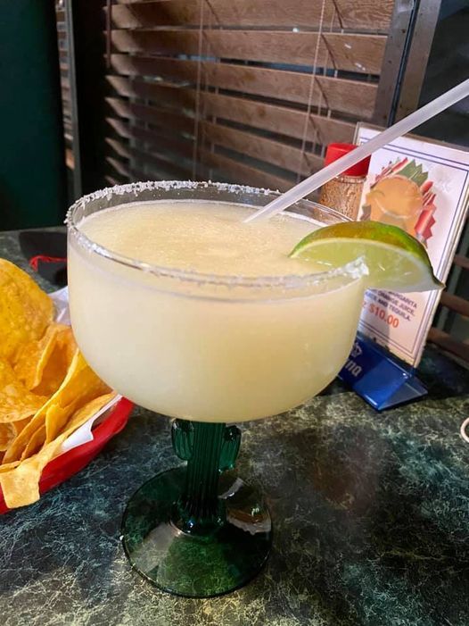 Margarita in a cactus-shaped glass with salt rim, lime wedge, and straw, on a table next to chips.