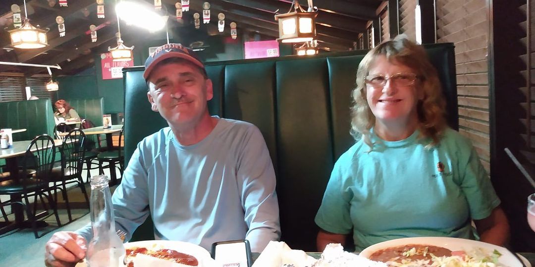 Two people seated at a table in a restaurant. The man wears a baseball cap, and the woman has glasses. Both are smiling.