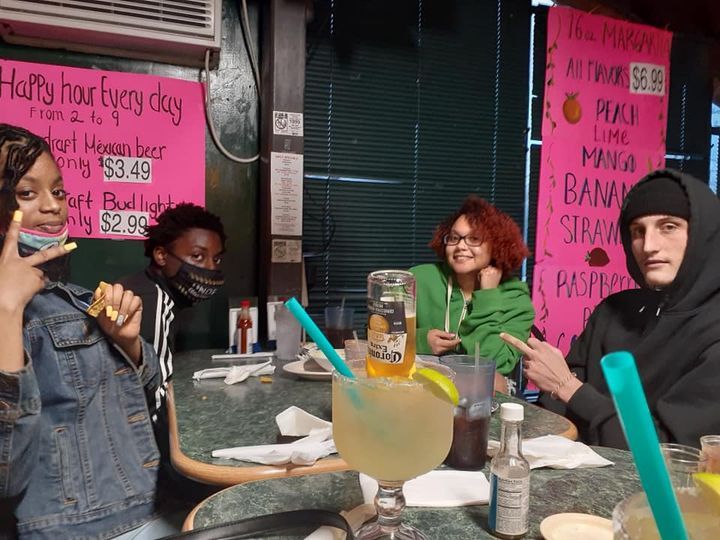 Four people sit at a table in a bar. One gives a peace sign, another points. Margaritas are visible.