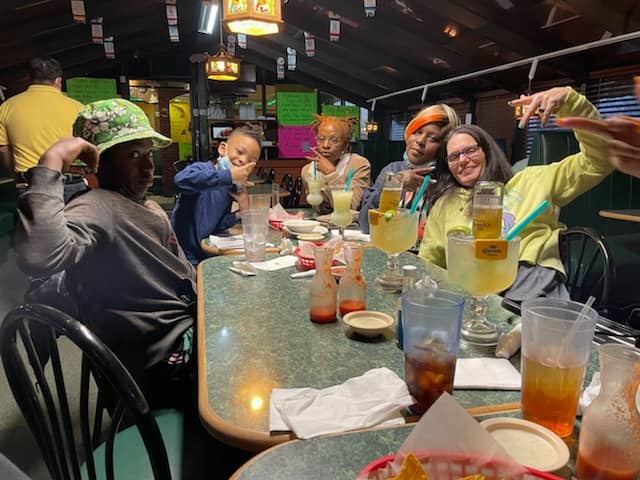 Group of people at a restaurant table with large drinks, some smiling and holding up hands.