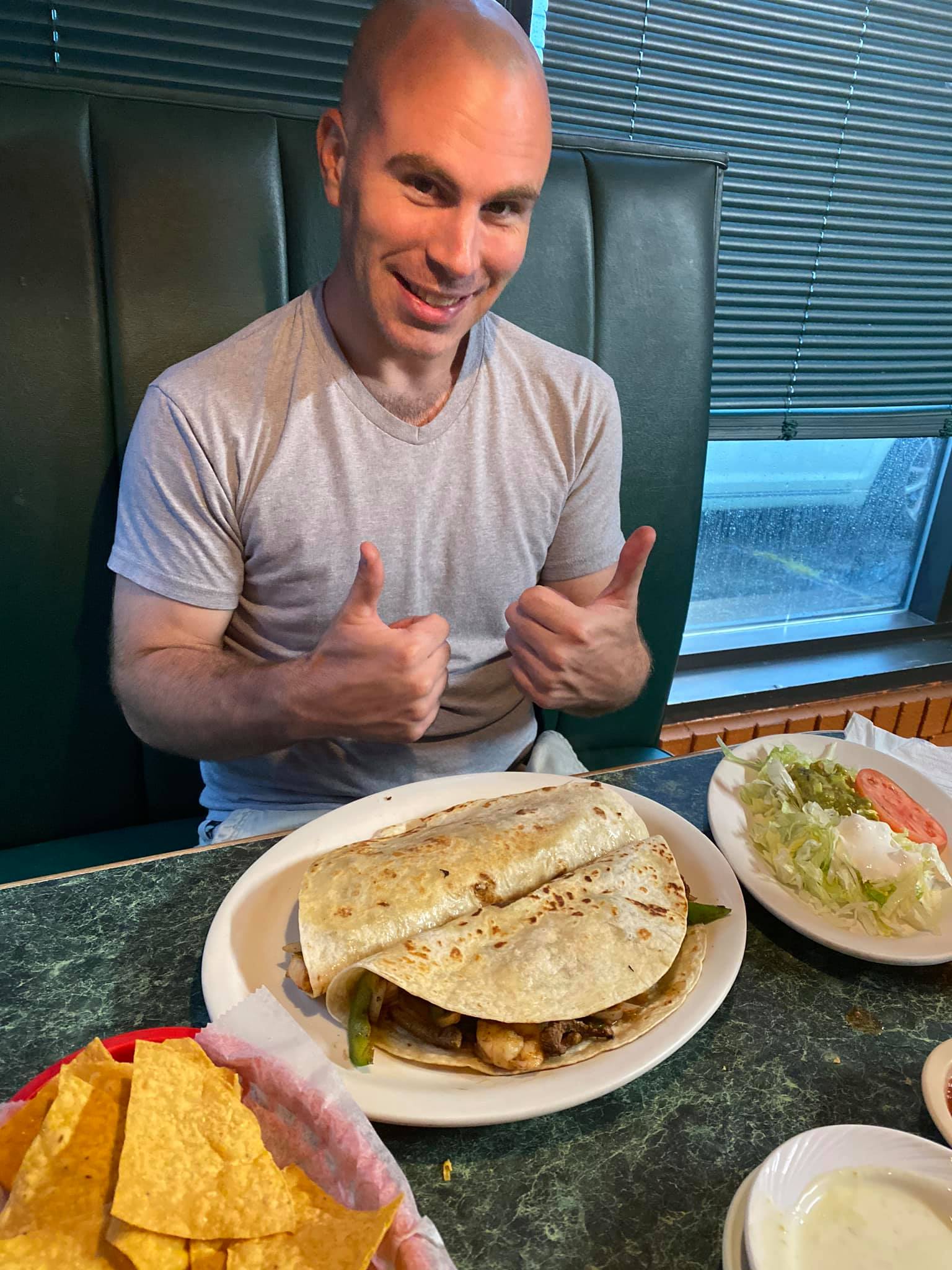 Man giving thumbs up at a restaurant, with a quesadilla, chips, and side dish on the table.