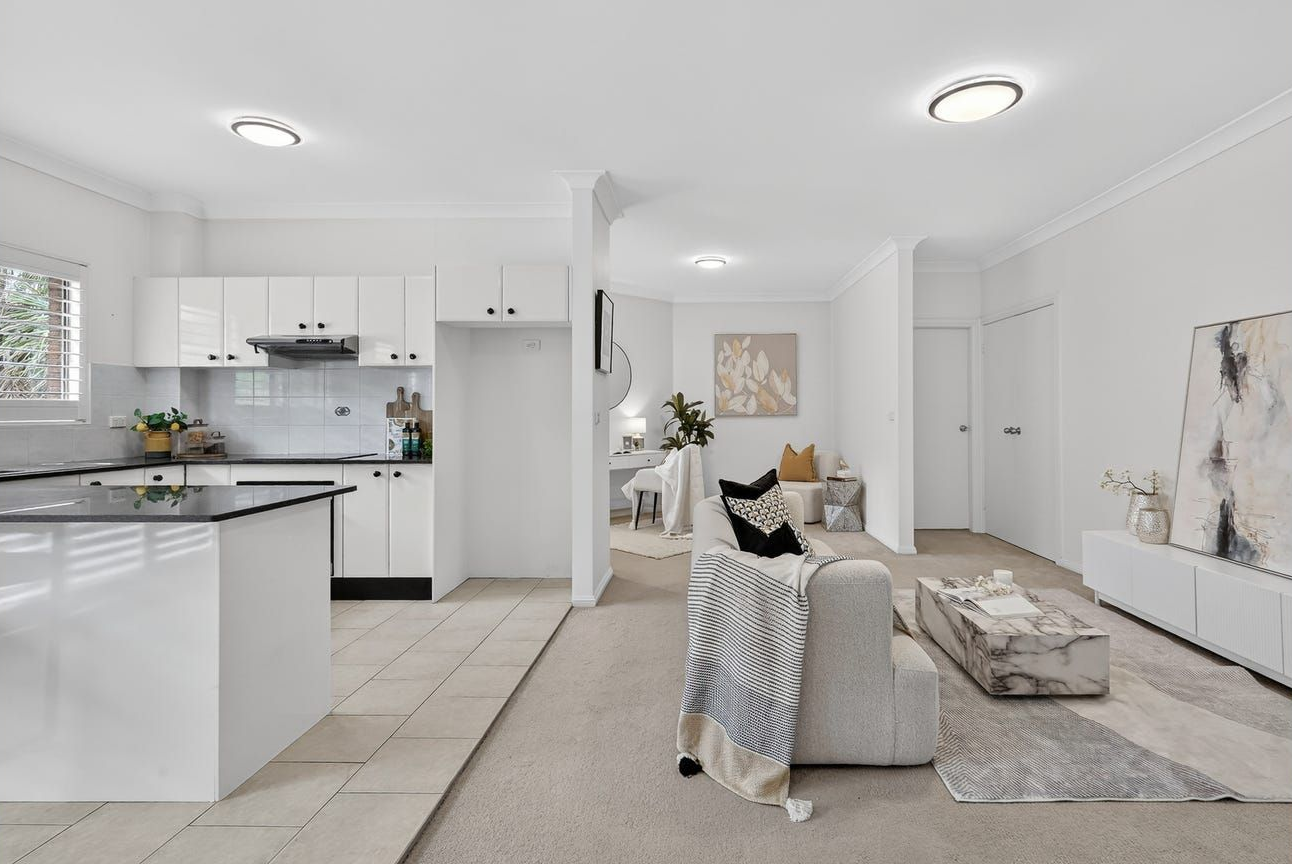 Modern open-plan kitchen with white cabinetry leading into a carpeted living area featuring a neutral sofa and artwork