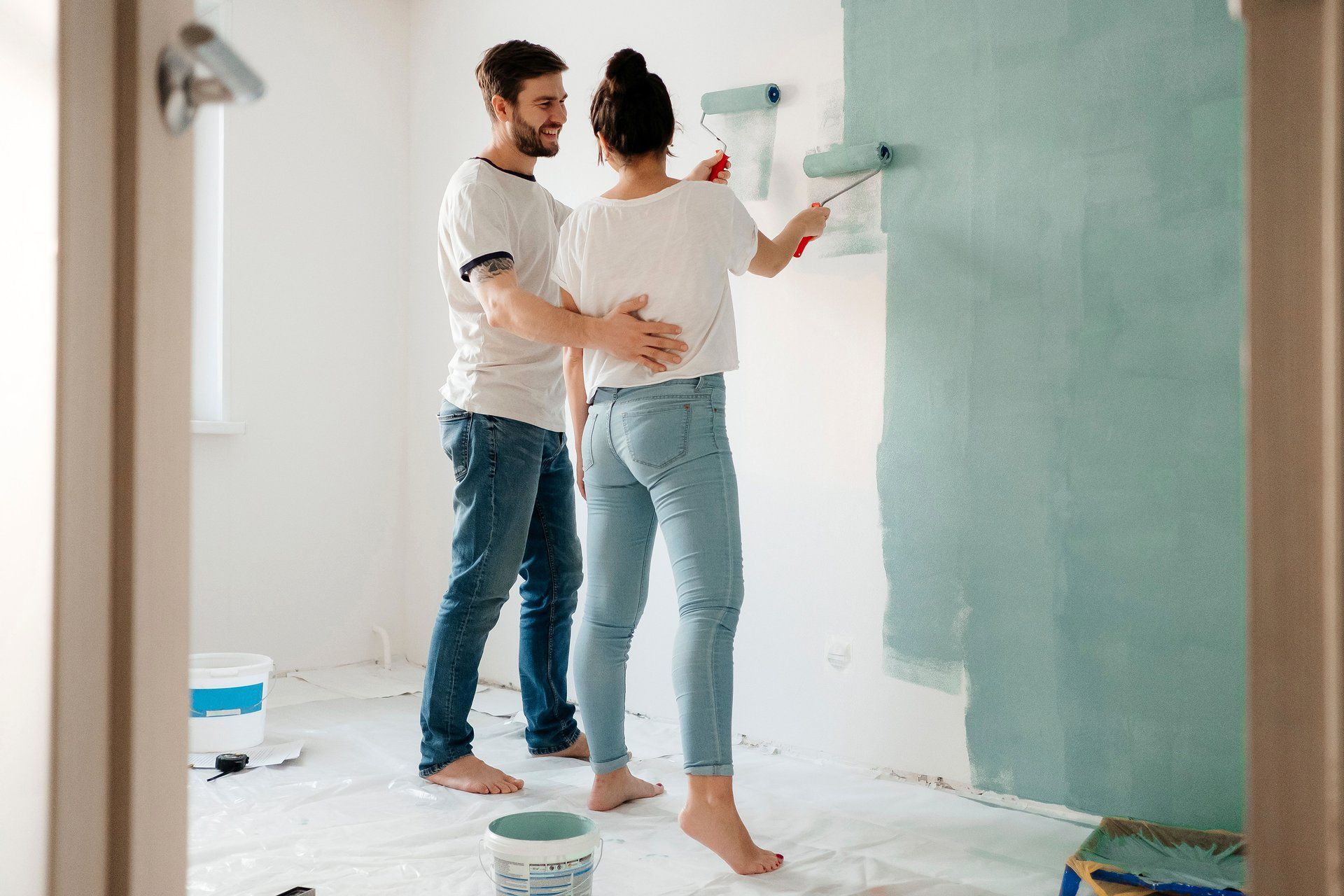 Couple painting a wall together in a bright room, with paint cans and a roller nearby.
