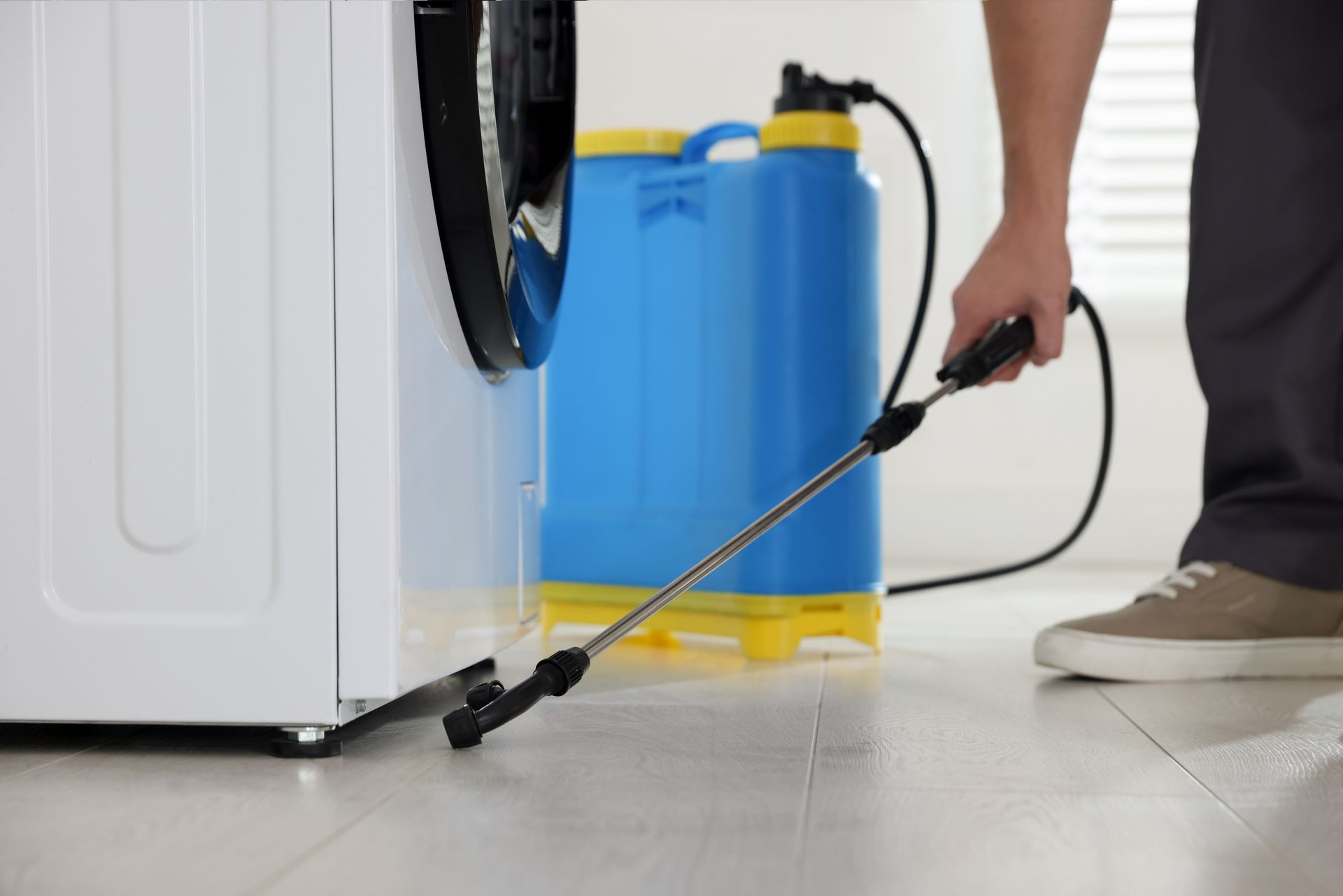 Pest control technician spraying treatment around a household appliance with a backpack sprayer.