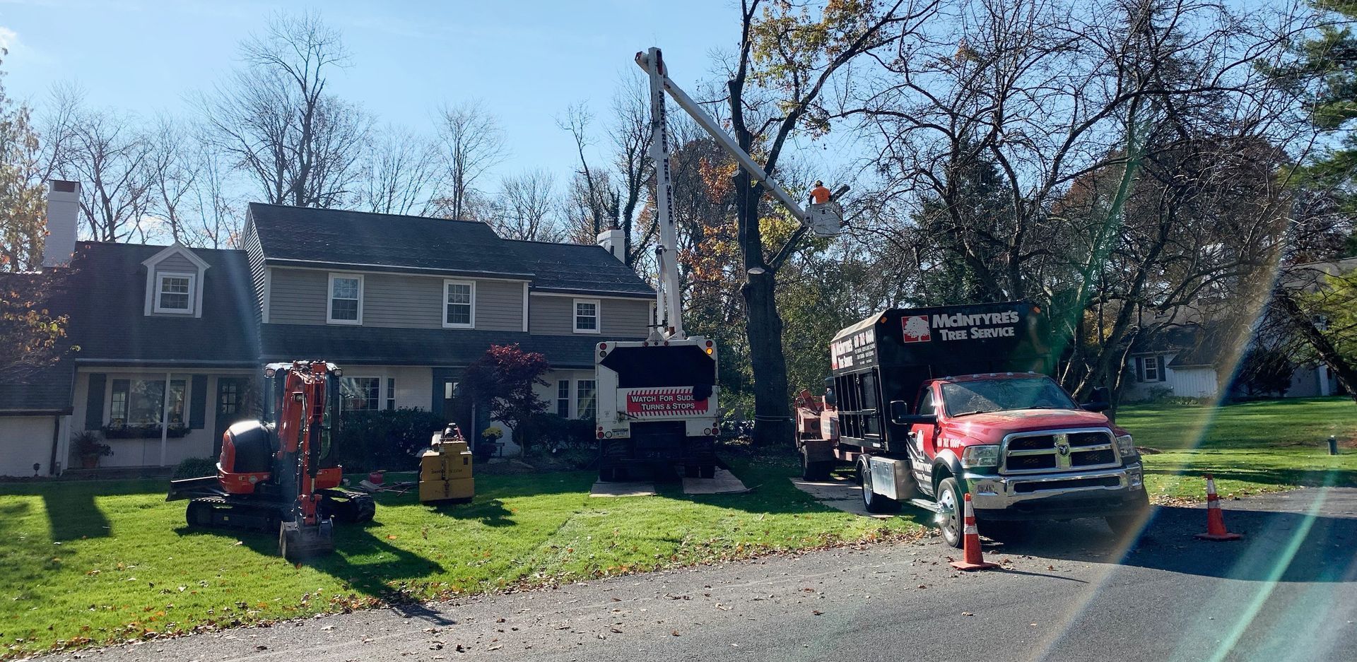 Tree service removing tree branches near a house, with equipment including a lift truck and excavator.