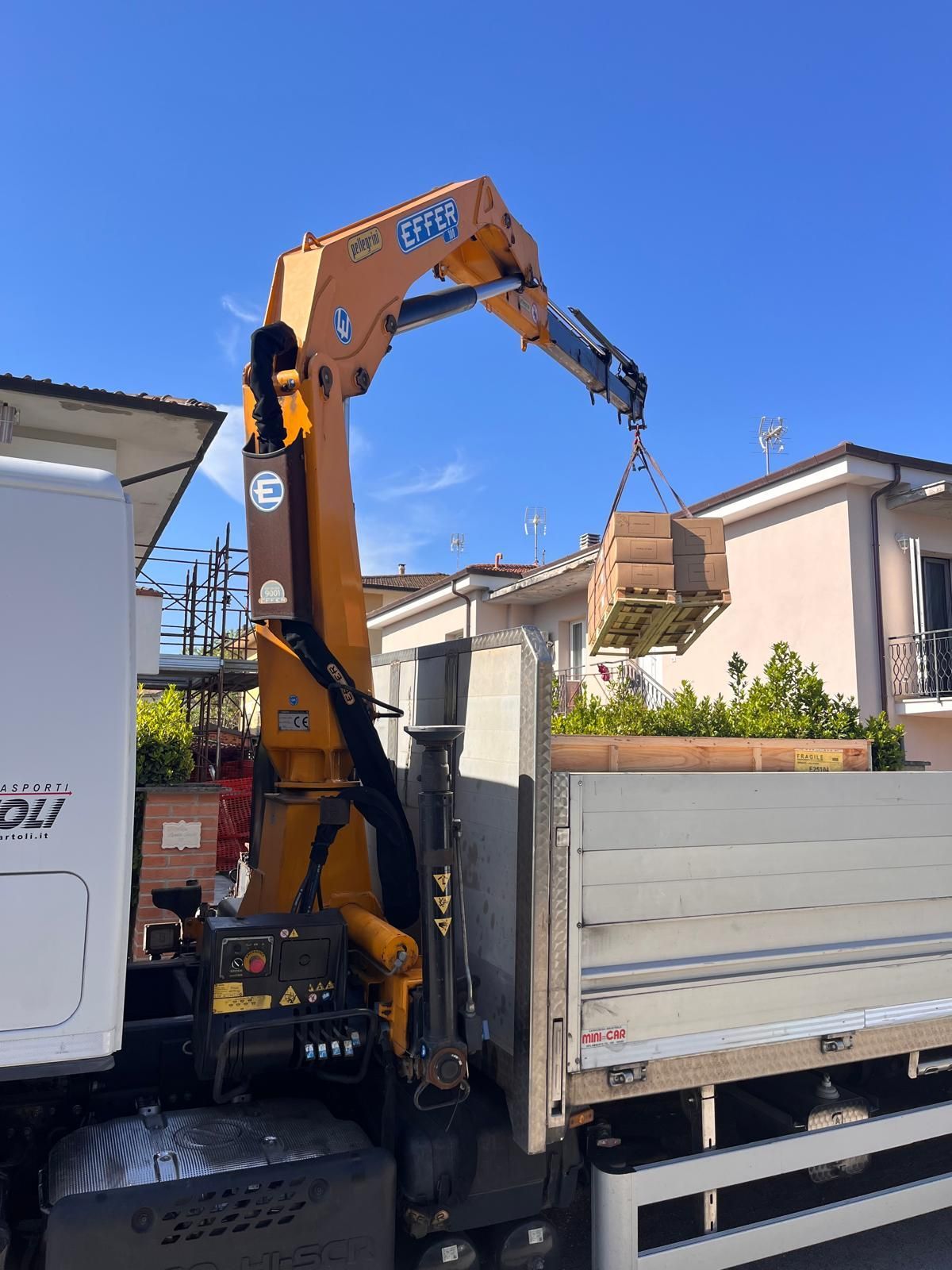 Un camion con gru arancione solleva detriti sopra un autocarro ribaltabile bianco in una strada residenziale sotto un cielo azzurro e limpido.