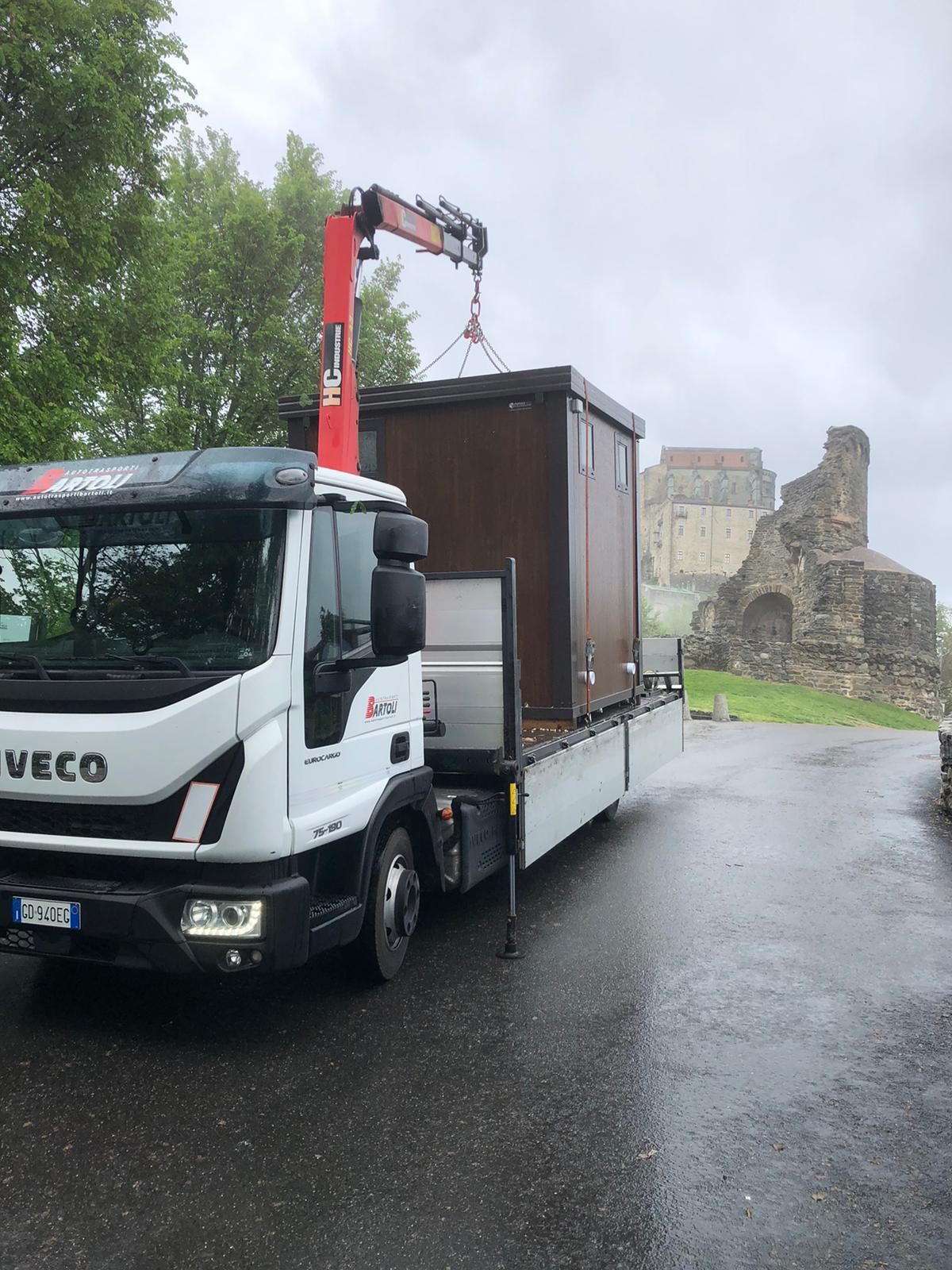 Un camion con gru arancione solleva detriti sopra un autocarro ribaltabile bianco in una strada residenziale sotto un cielo azzurro e limpido.