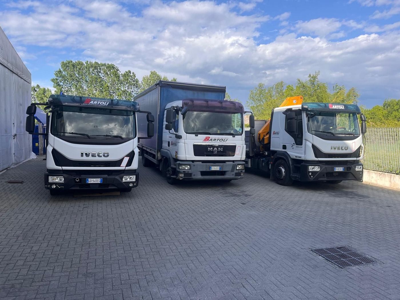 Tre camion Iveco parcheggiati in un piazzale, uno bianco e uno grigio, con alberi e cielo sullo sfondo.