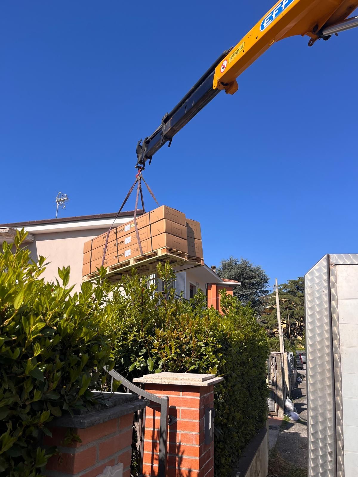 Una gru solleva fasci di mattoni sopra una casa in un soleggiato cortile residenziale.