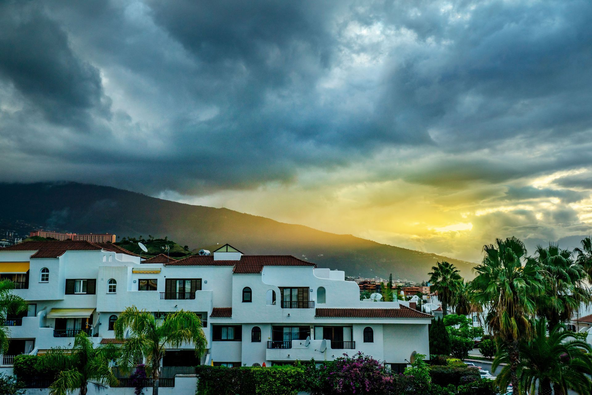 White villas beneath dark storm clouds with a glowing sunset over distant hills and palm trees