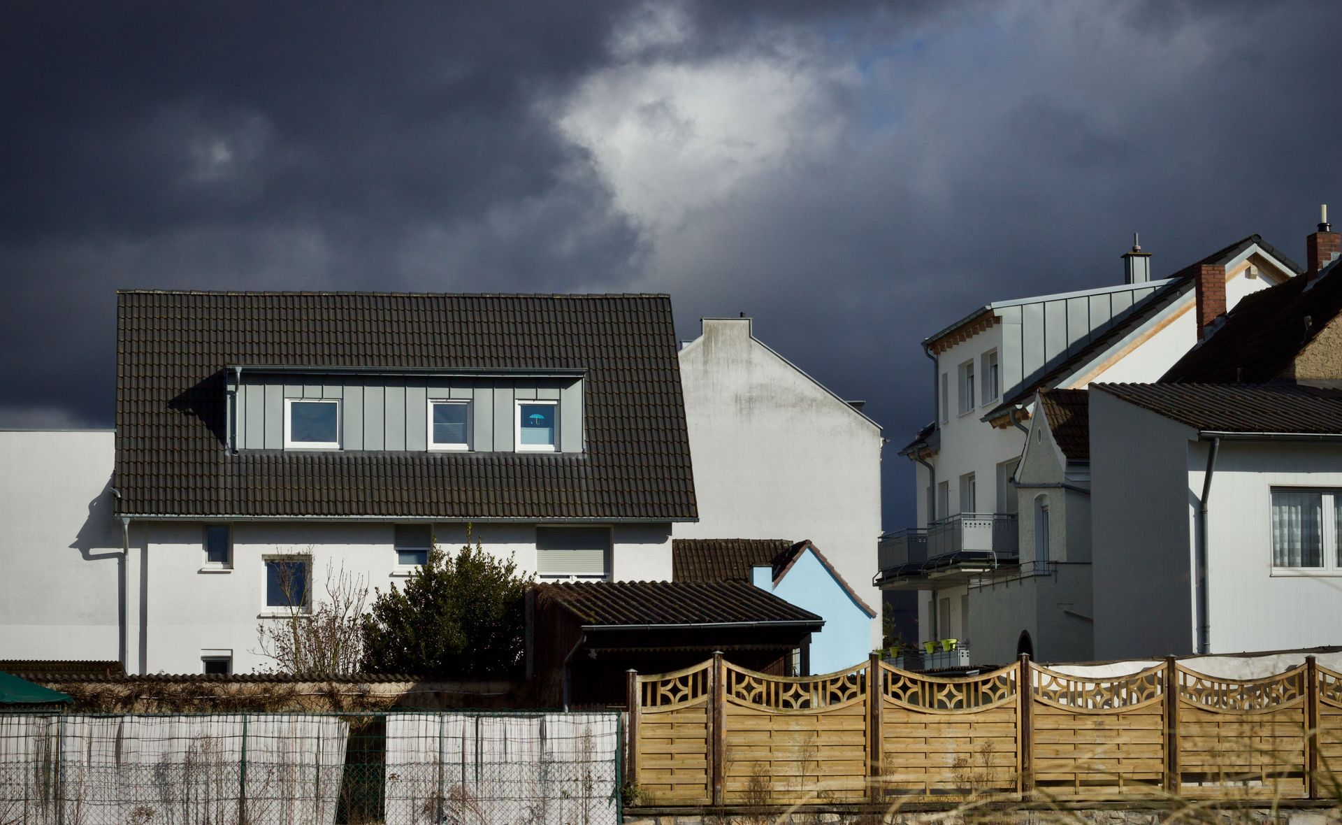 White houses with a dormer roof behind a wooden fence under dark storm clouds.