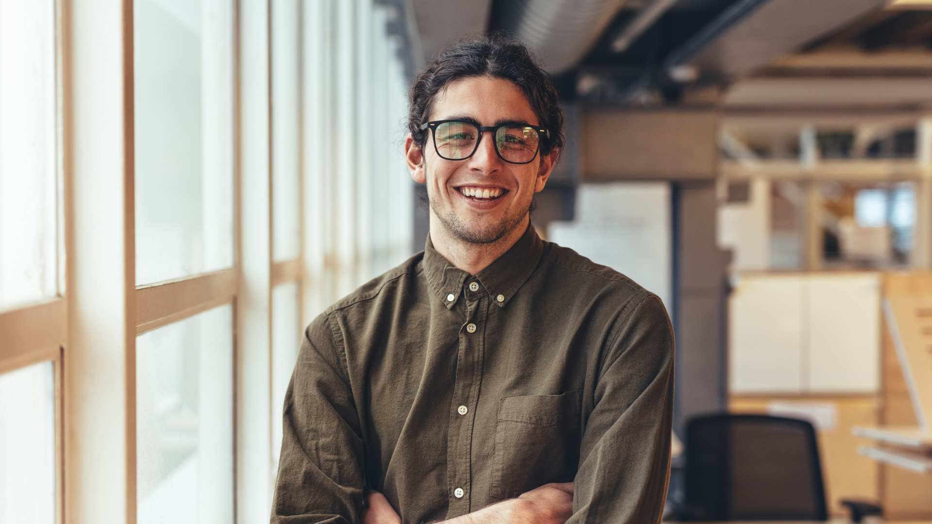 Person smiling in glasses and olive shirt, standing in a bright office with large windows.