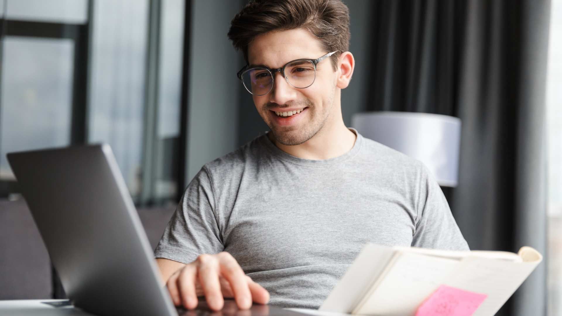 Smiling man using a laptop and notebook at a desk in a bright office